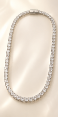 A tennis-style necklace paved with clear round stones in a silver-toned setting, arranged in an oval shape on a softly lit cream background. A small engraved logo plaque is visible at the clasp.