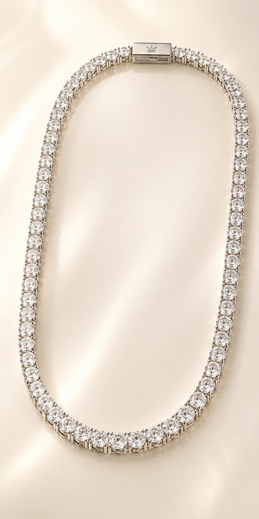 A tennis-style necklace paved with clear round stones in a silver-toned setting, arranged in an oval shape on a softly lit cream background. A small engraved logo plaque is visible at the clasp.